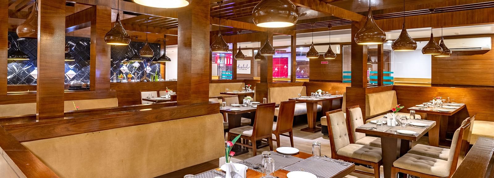 Warmly lit restaurant at Diamond Palace Lords Plaza with wooden beams, set tables, and booth seating.