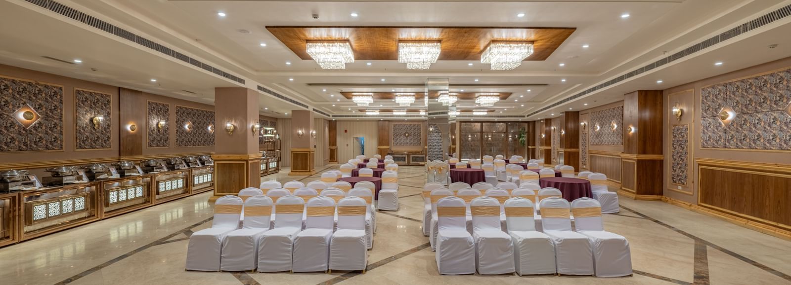 Large banquet hall at Hotel Highway King, Bagru, with rows of chairs, decorative ceiling lights, and mirrored wall panels.