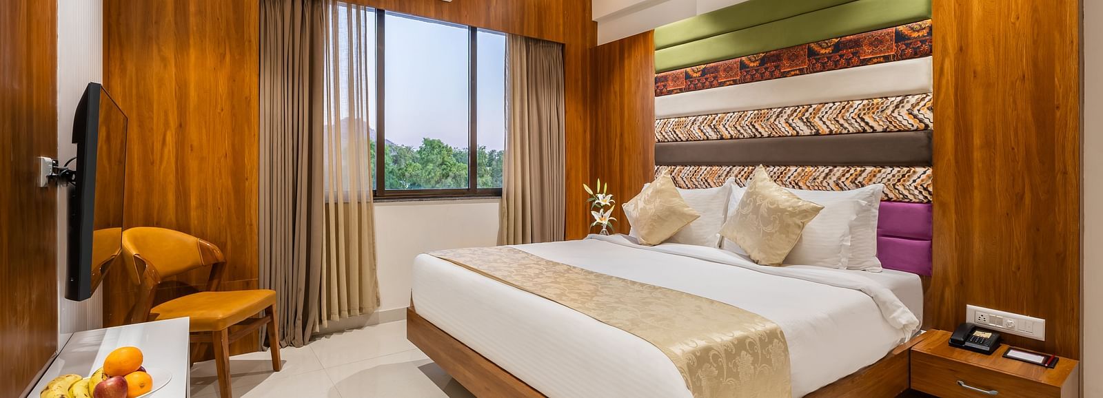 A comfortable king-size bed with plush pillows and a throw dominates a hotel room with a TV and a cushioned chair - Lords Eco Inn Ambaji.