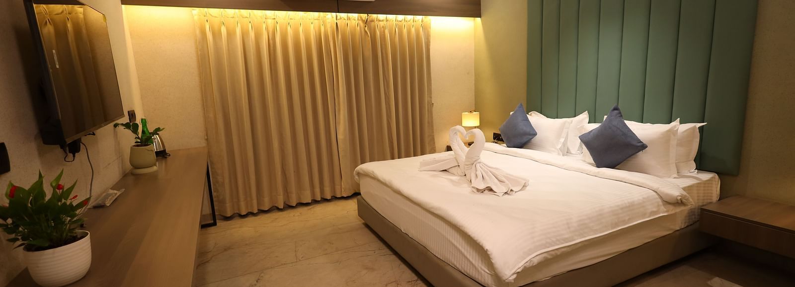 A well lit elegant room featuring a king size bed with decorative towels and pillows, bedside lamps, illuminated draped curtains and a mounted T.V. above an elongated desk with potted plants at Lords Eco Inn Navsari.