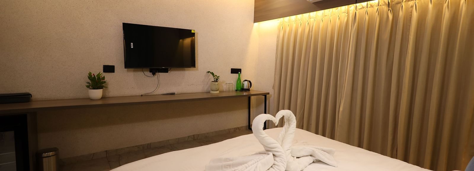 The image shows a hotel room with a bed featuring swan-shaped towel art, a television, a desk, and curtains - Lords Eco Inn Navsari.
