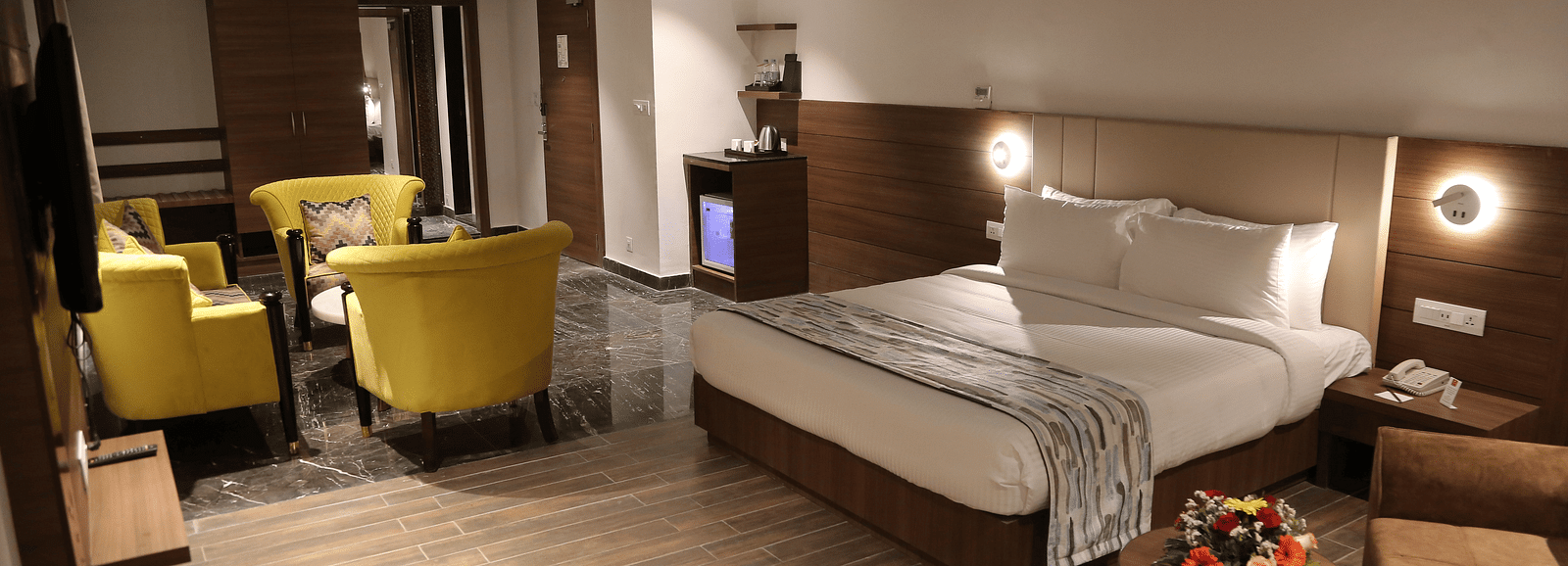 A spacious hotel room features a neatly made bed, a seating area with yellow armchairs, and modern decor under soft lighting.