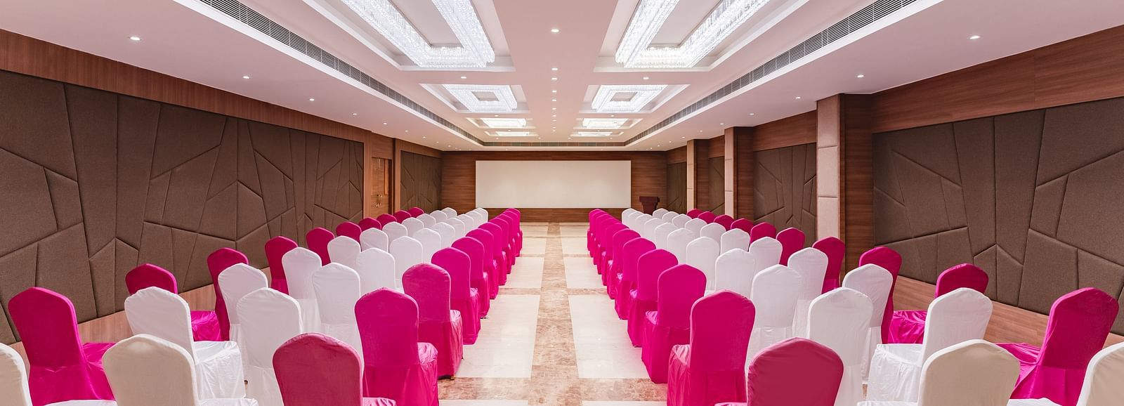 A large banquet hall at Lords Inn Udaipur is arranged with rows of tables and chairs draped in white and vibrant pink fabric, facing a stage.