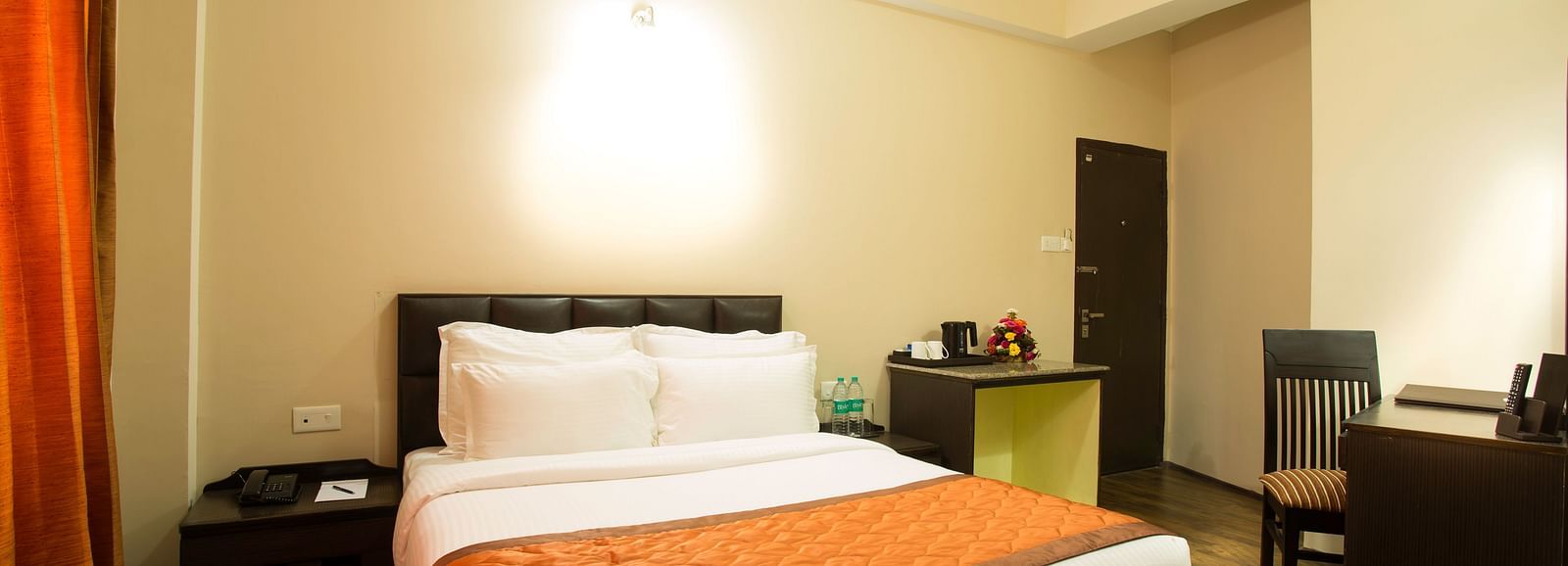 A room at New Orchid Lords Inn Gangtok contains a bed with pillows, a bedside table with a phone and water, curtains, a desk with a chair, and a luggage rack.