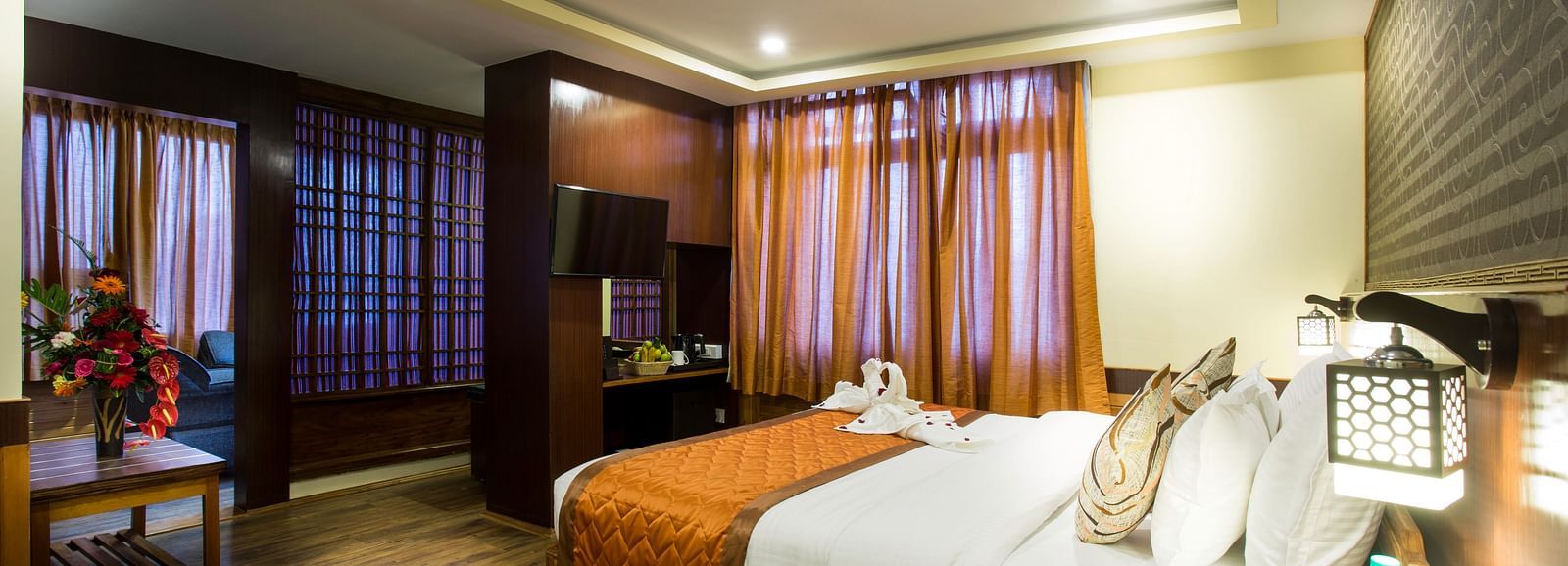 A room at New Orchid Lords Inn Gangtok contains a bed with pillows and folded towels, bedside tables with lamps and water, curtains, and a table with flowers.