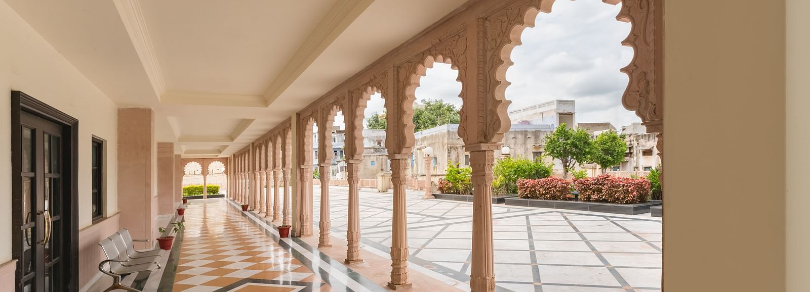 A covered outdoor corridor at Shri Vallabh Vilas Lords Plaza Nathdwara, with arched stone columns and checkered flooring, opening to a garden area.