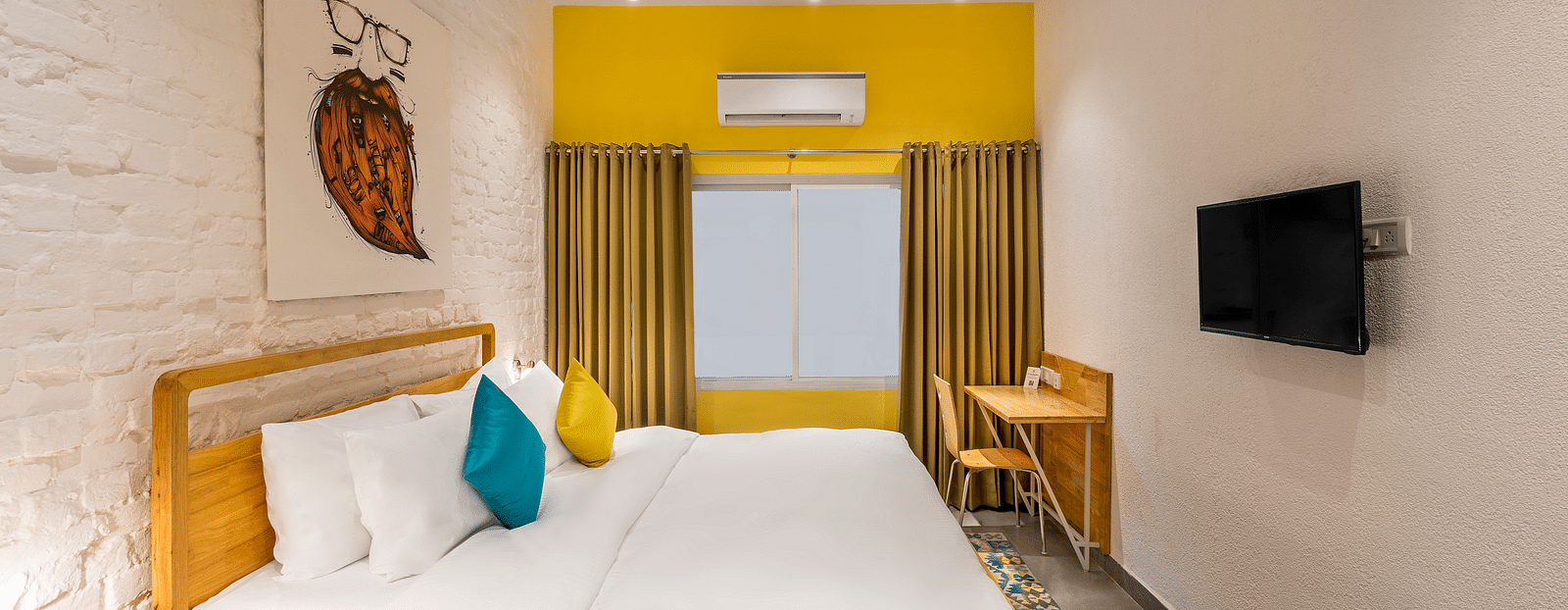An side angle interior image of the standard room at Bedzzz Varanasi showcases a spacious layout, featuring a comfortable double bed, a cozy floor mat, a stylish study table and chair, a charming coffee table, elegant curtains, an air conditioner for a ref
