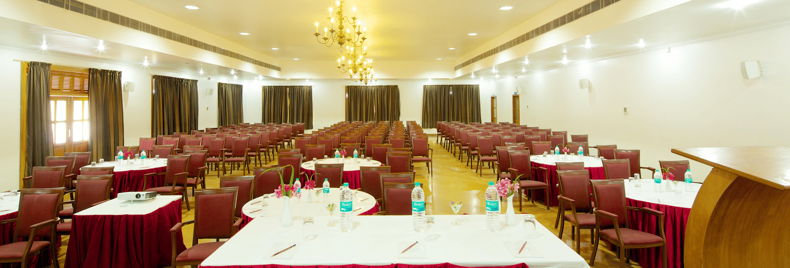 A large meeting room arranged with multiple tables covered in white tablecloths with red valances and surrounded by chairs | Le Pondy