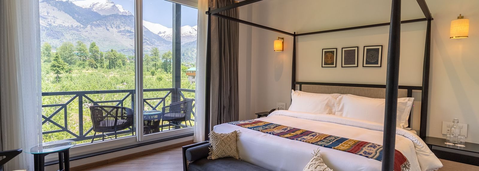 Spacious room with a large wooden bed, cushioned seating at the foot, and a glass door opening to a balcony with a serene nature view at Nivasana Manali.