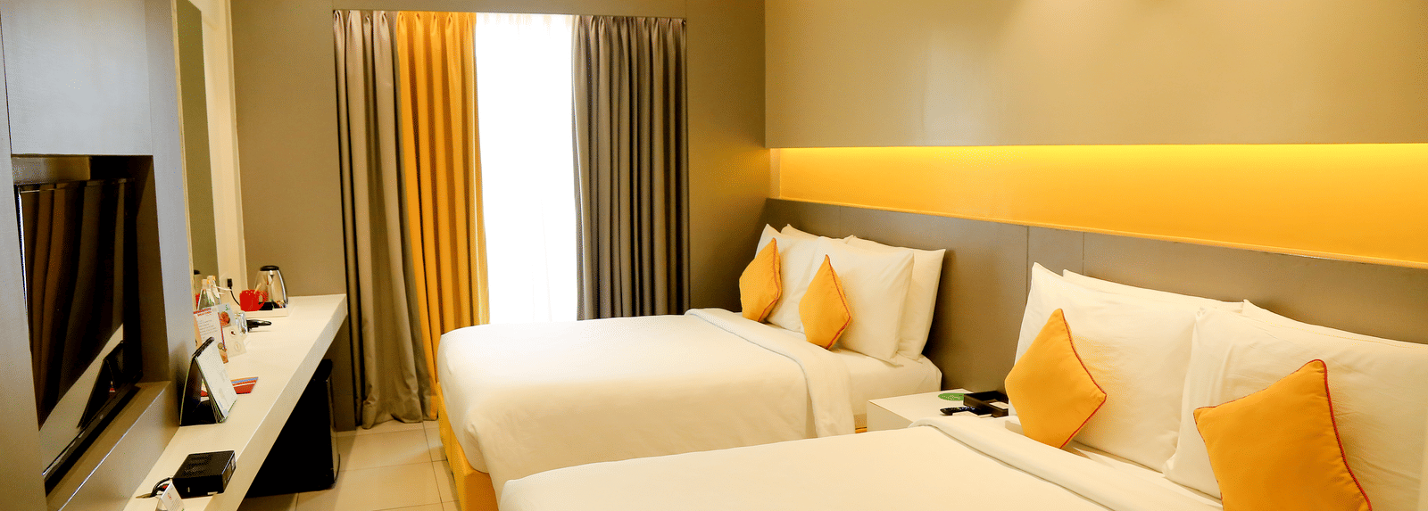 A spacious room with twin beds featuring yellow cushions and headboards, large windows with curtains, and a wall-mounted TV - Zibe Coimbatore by GRT Hotels