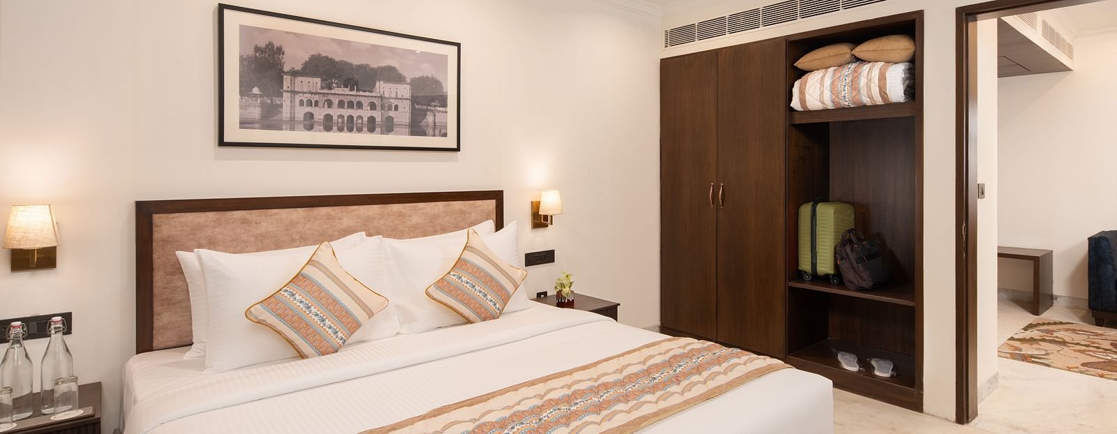 An image of a bedroom with king size bed, photo frame at the top, bedside table with lamp on either side, and a door giving me a sneak peak of  another room - Brij Eternity, Vrindavan