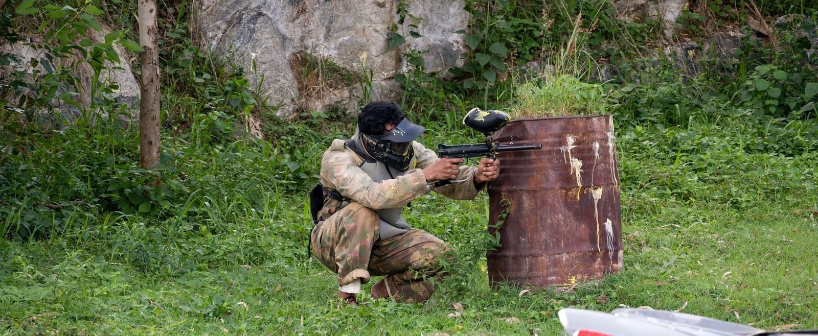Two individuals are engaged in a paintball game in a natural setting, with one person taking cover behind a wooden barrier | RD's Nature Retreat, Bangalore