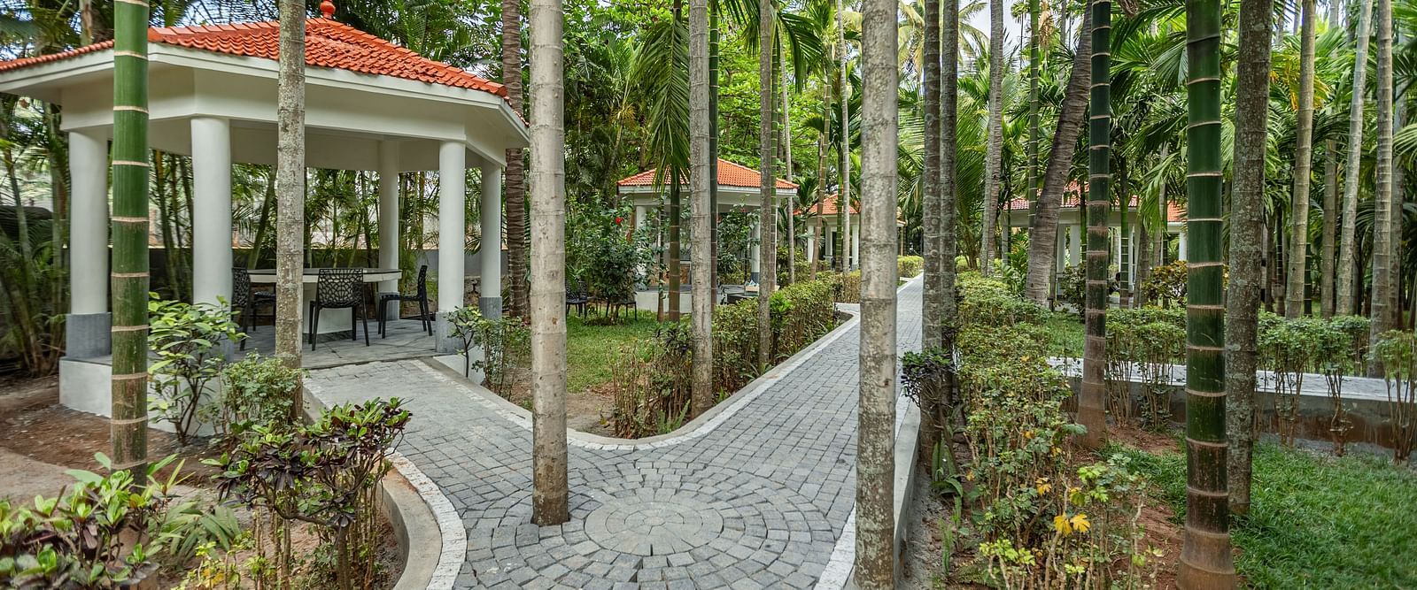 A white gazebo with an orange roof and a paved pathway winding through palm trees and greenery - Mango Hill Resort, Coimbatore