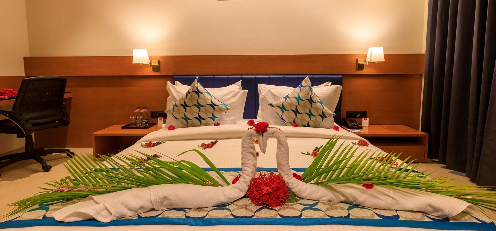 A decorated double bed in a warmly lit room at Lords Eco Inn Apple Gate, featuring towel swans and floral arrangements.