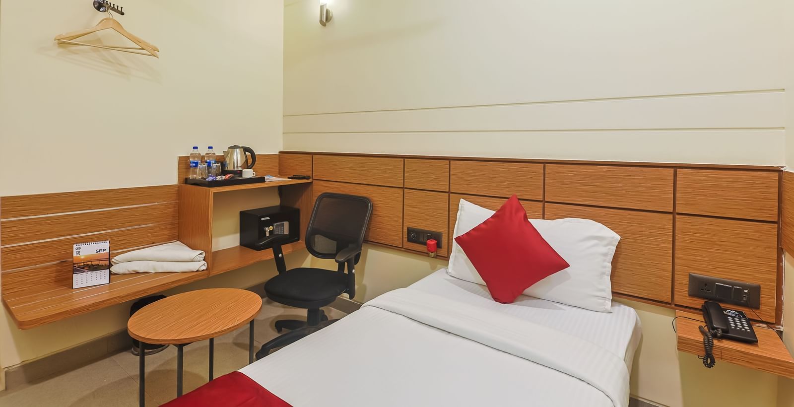 Bedroom with single bed, red runner, pillow, chair, desk, hanger and air conditioner at Max Hotel Prayagraj.
