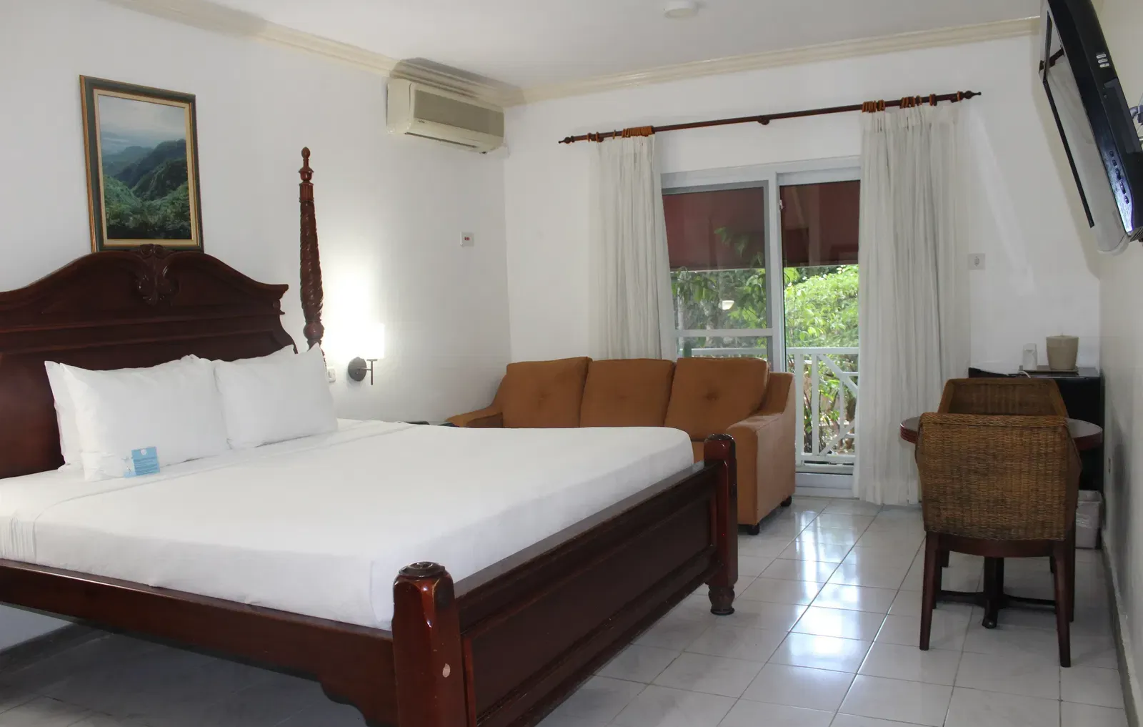 A hotel room features a large wooden bed with white linens, a dark dresser with a television, and a seating area with a couch | Deluxe King Room | Altamont Court, Jamaica