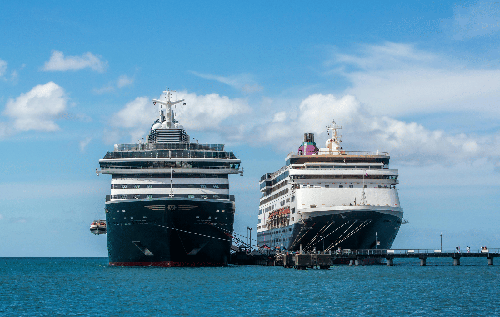 A large cruise ship docked alongside a smaller vessel at a busy port terminal, with a clear open sky above and calm harbour waters surrounding the pier and docking facilities.