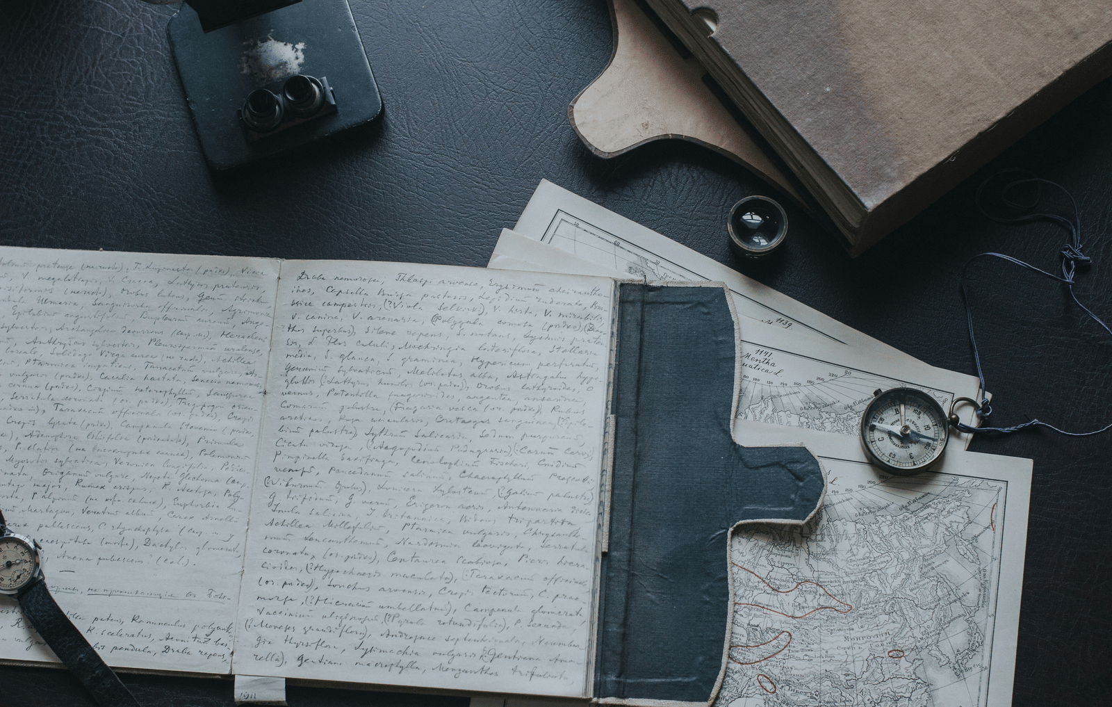 A flat-lay arrangement of antique-style maps, a compass, and a leather notebook.