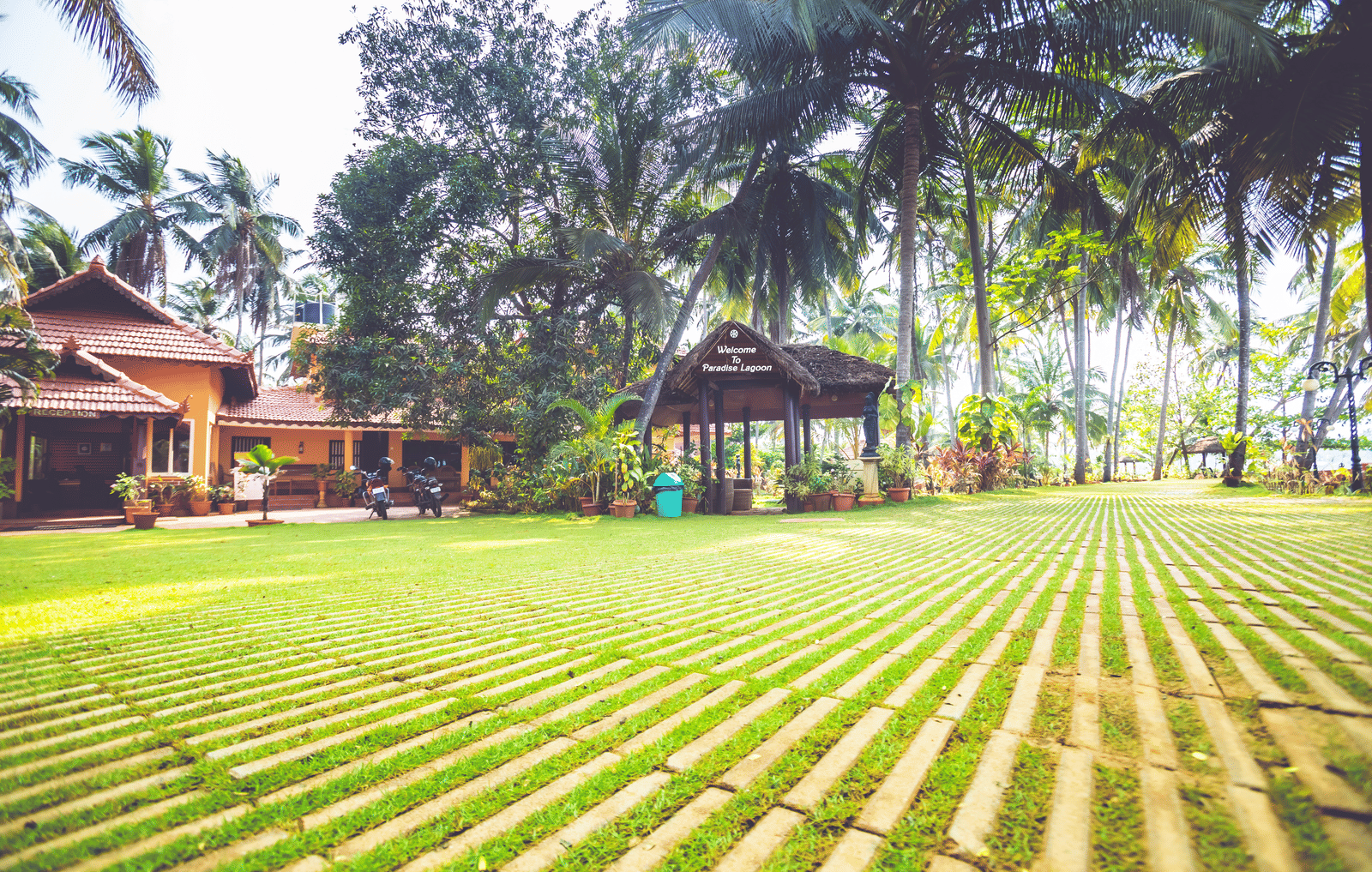 Lawn with palm trees, tiled pathways, and red-roofed building surrounded by greenery at Paradise Lagoon Resort, Udupi.