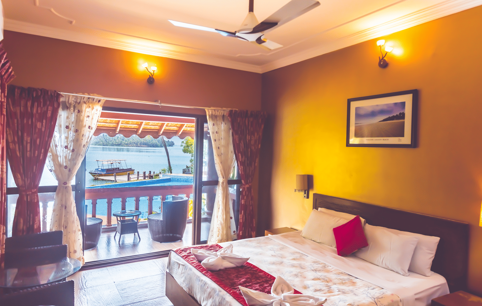 Room with a queen size bed, ceiling fan, framed picture, and view of the river through the window at Paradise Lagoon Resort, Udupi.