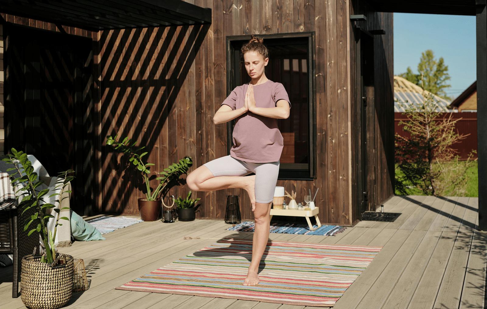 A woman practises yoga on a striped mat in an outdoor wooden deck area, surrounded by potted plants and sunlight filtering through a slatted roof.