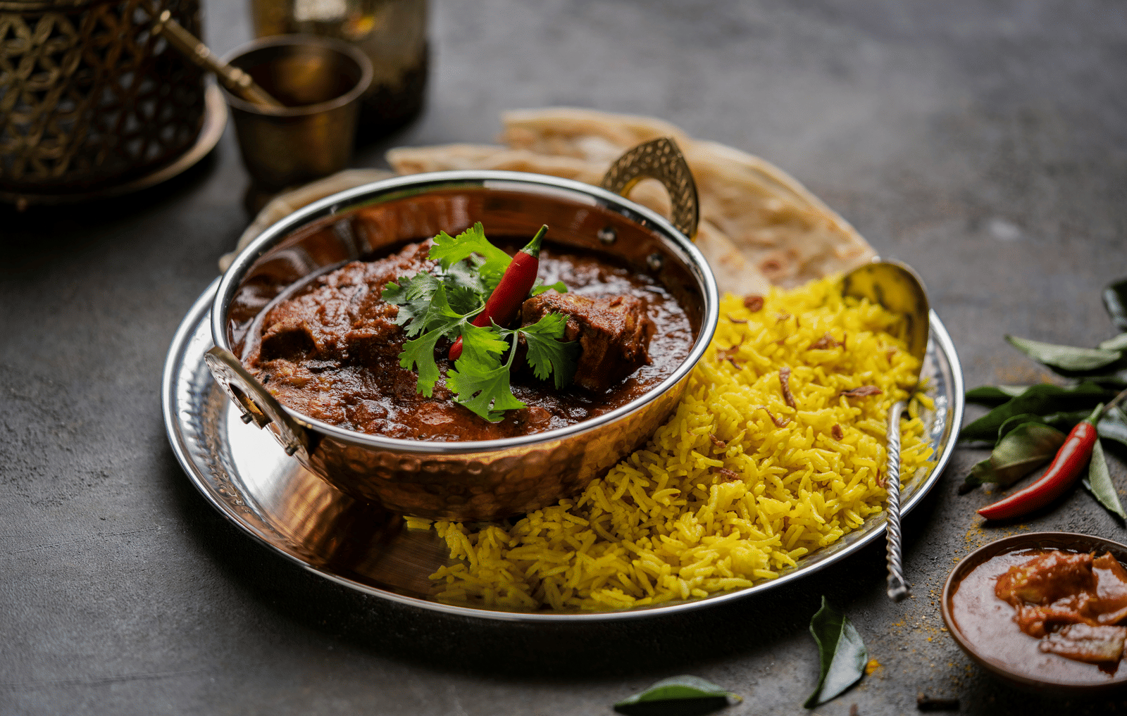 Bowl of spicy curry served with yellow rice and garnished with herbs on a dark table.