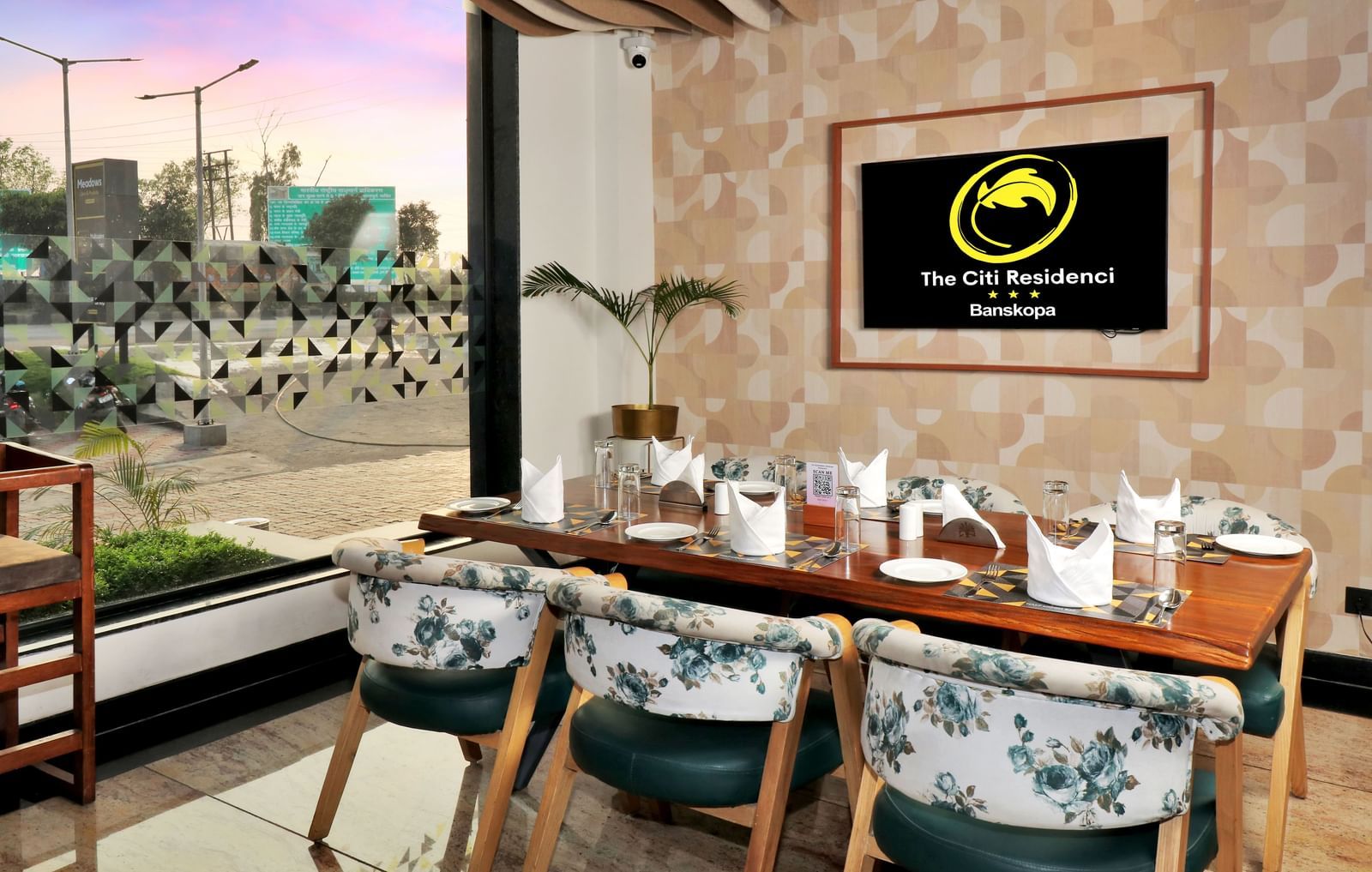A restaurant dining area with a long wooden table set for guests, surrounded by chairs with floral upholstery, and a framed logo on the textured wall | The Citi Residenci Hotel Banskopa
