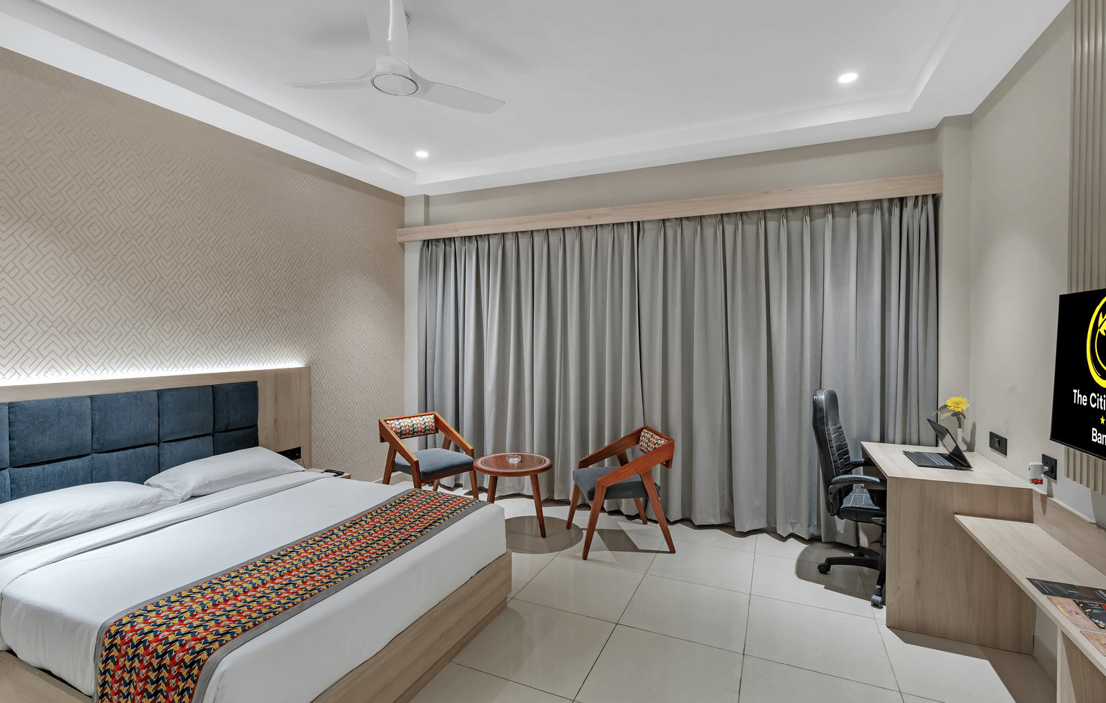 A well-lit hotel room with a queen size bed featuring a colourful patterned throw, a seating area with two chairs and a small table, a work desk with a chair, and a television | The Citi Residenci Hotel Banskopa