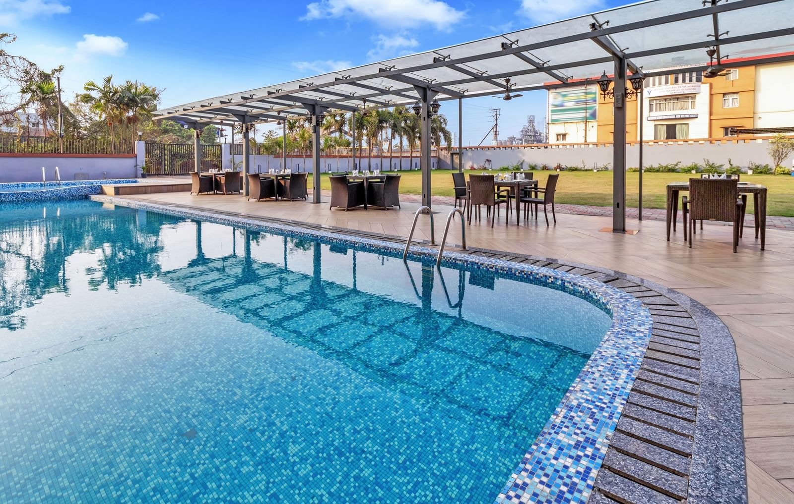 A sparkling blue outdoor swimming pool with mosaic tiling along the edge, surrounded by a paved area and covered dining tables under a clear sky with scattered clouds  | The Citi Residenci Hotel Banskopa