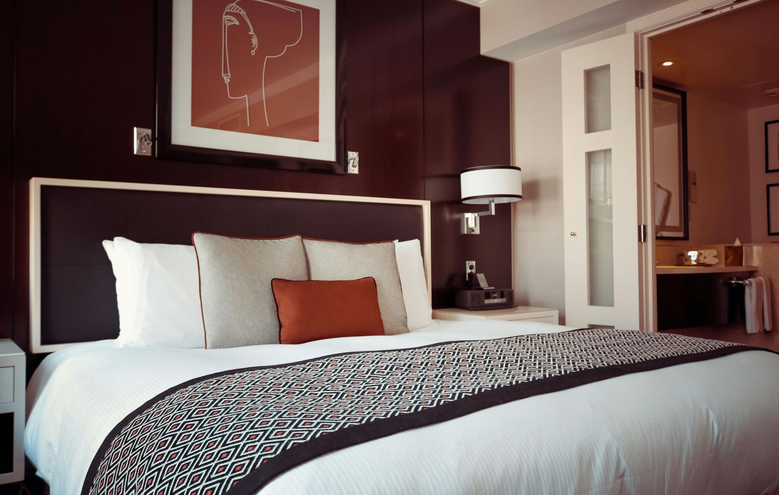 Elegantly decorated hotel bedroom with a large bed, multiple pillows, a patterned bed runner, artwork above the headboard, and a glimpse into an adjoining room.