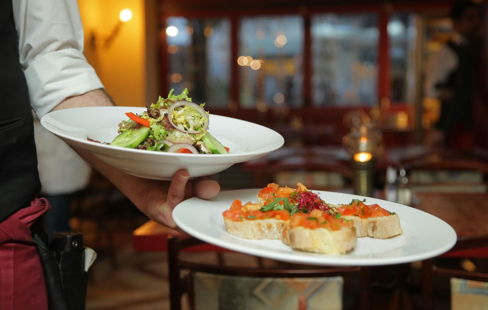 A waiter in a white shirt and black vest carefully carries a plate of salad and a plate of bruschetta in a warmly lit restaurant setting.