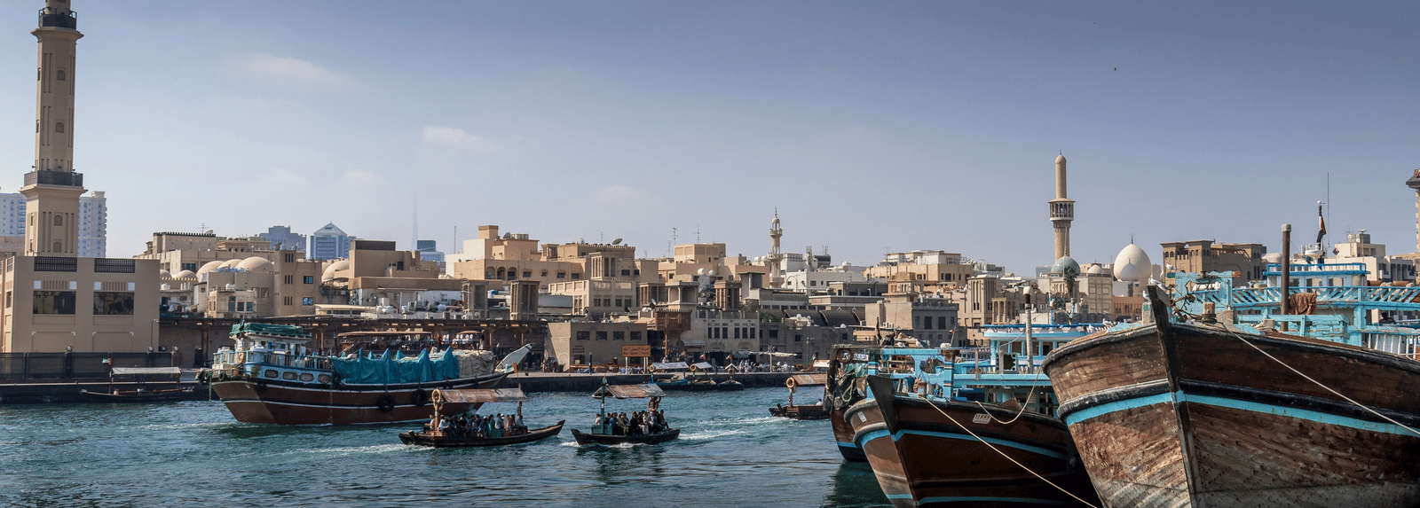 Traditional wooden abra boats docked along a waterfront with city buildings and a mosque visible in the background.