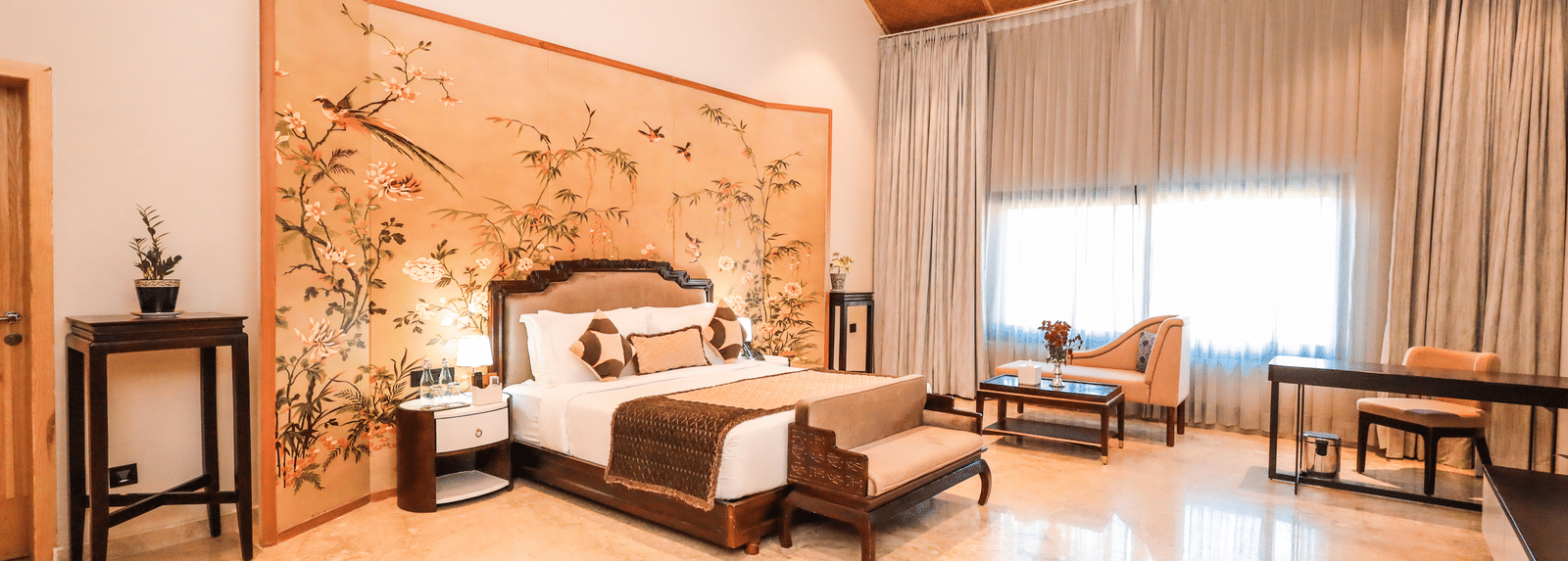 A room shows a bed with side tables, a bench at the foot of the bed, a seating area with chairs and a large window with curtains at Ananta Spa & Resort, Jaipur.