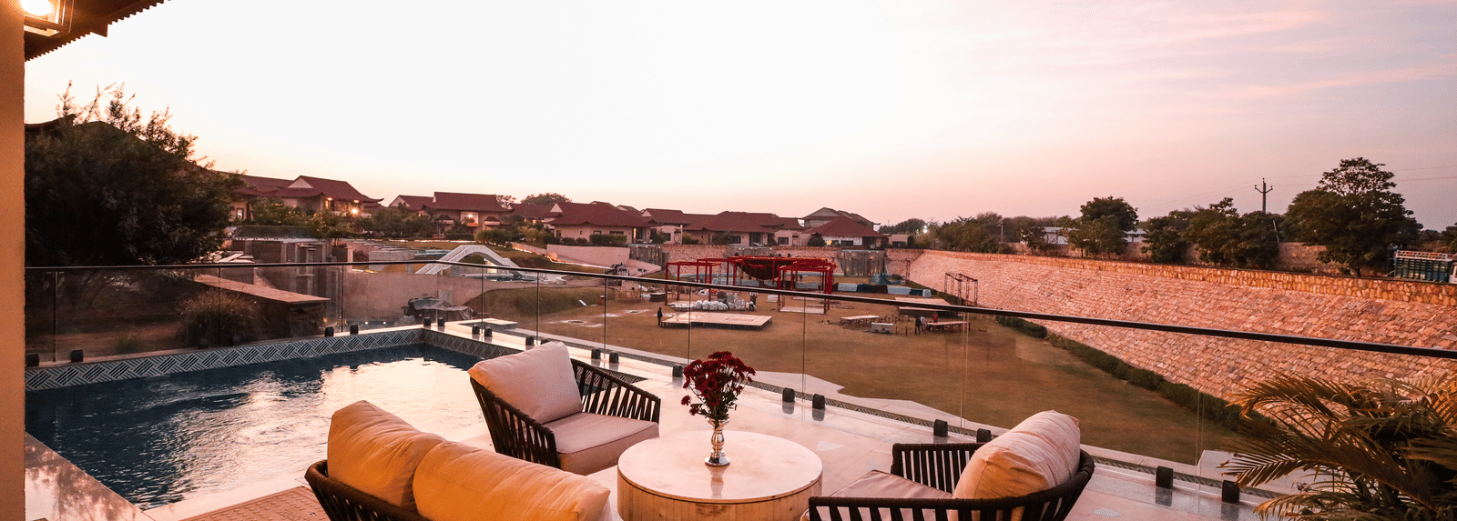 An outdoor space shows lounge chairs and tables beside a pool with landscape views in the distance at Ananta Spa & Resort, Jaipur.