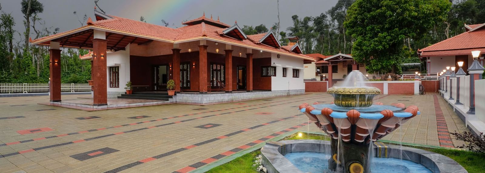The traditional facade of Bhuthanakadu Retreat under a vibrant rainbow, featuring a fountain in a garden and a colorful patterned walkway leading to the main buildings.