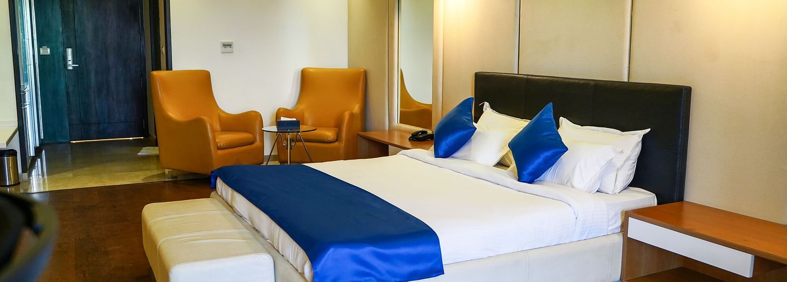 A stylish hotel room at BNGV Grandeur Hotel and Banquets, equipped with a bed featuring a dark headboard and blue accents, complemented by two vibrant orange armchairs.