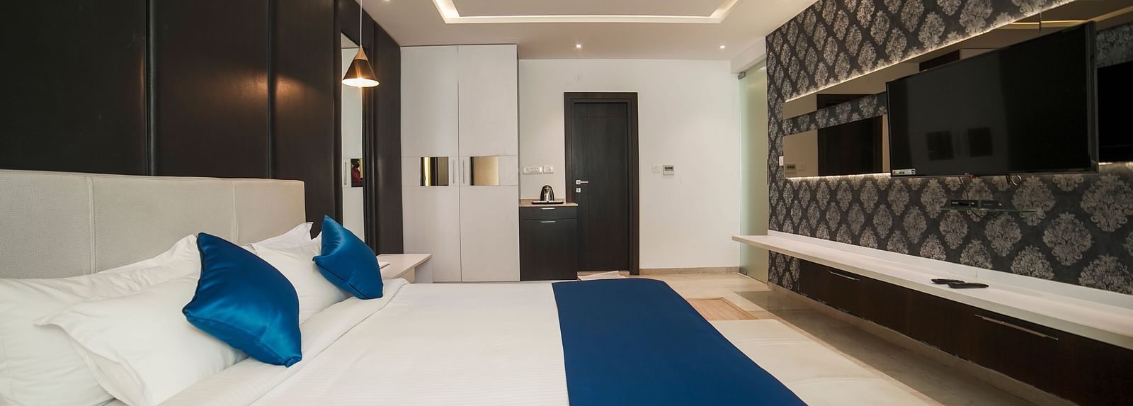A modern hotel room at BNGV Grandeur Hotel and Banquets, featuring a comfortable bed with a blue bed runner, clean white linens, and sleek furnishings.