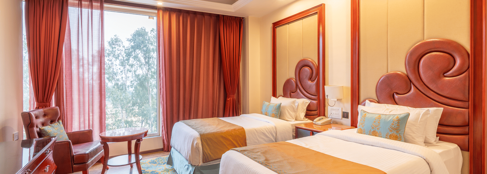 Twin bedroom with rich wooden ceiling beams, gold and blue accents, 2 large beds, and a view through the window at Hotel Hukam's Lalit Mahal.