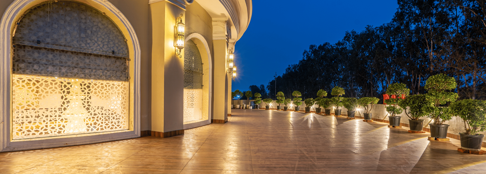 Curved, illuminated walkway at night with decorative wall panels, a polished floor, and potted plants lining the path at Hotel Hukam's Lalit Mahal.
