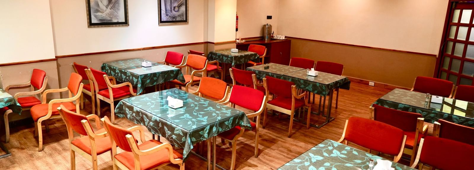 An empty dining room with multiple tables, patterned tablecloths, and several upholstered chairs at Infiniti Hotel, Indore.