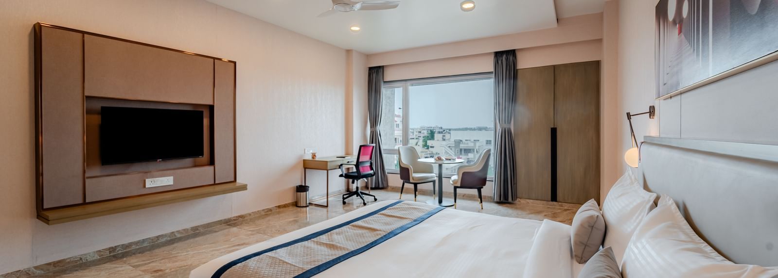  A wide angle shot of a hotel bedroom with a large bed, TV, and a desk, well-lit | Premium Room | Pravasa Hotel, Vijayapura