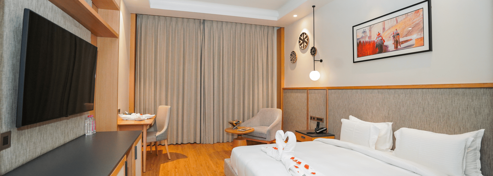 A clean and modern hotel room with a large bed, a TV, and a picture above the bed. The room has wooden floors and a ceiling fan.