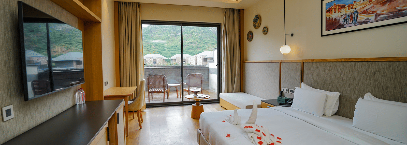 A well-lit hotel room with a large bed, a television, and a sliding glass door leading to a patio. There are rose petals on the bed.