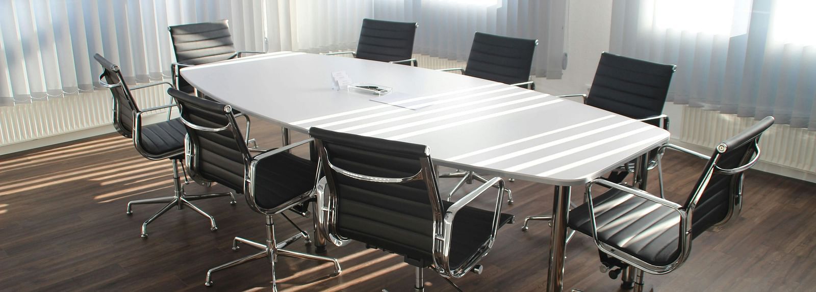 A close up shot of a brightly lit boardroom featuring a white long table along with office chairs surrounding it.