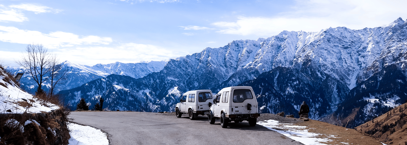 Two off-road vehicles parked along a mountain road overlooking a panoramic range of snow-covered Himalayan peaks.