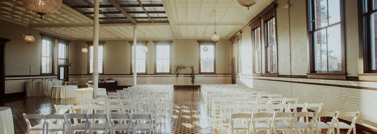 A hall arranged with chairs in a row, cloth covered tables, and chandeliers in front of large windows.