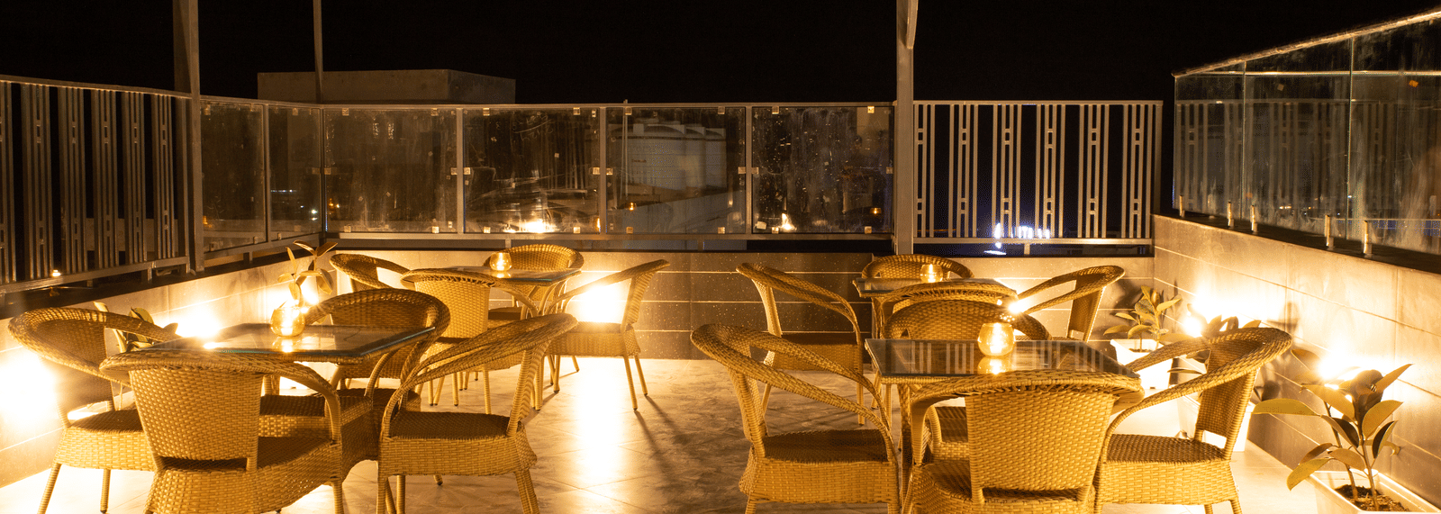 An outdoor rooftop cafe at Perfectstayz Premium Laxmi Heritage with woven chairs and tables on a patio, warmly lit under a dark sky, enclosed by a glass railing.