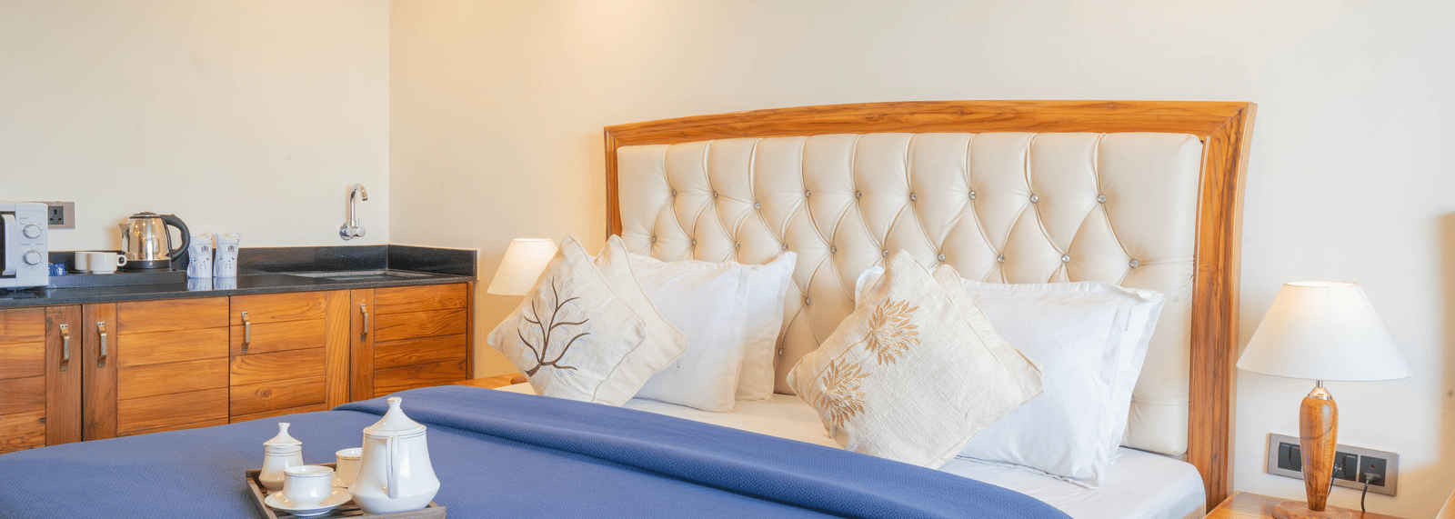 Close up of the king size bed in the Junior Suite of Perfectstayz Premium Mokshada Suites, Mussoorie, highlighting the plush cushions, tea & coffee maker placed on top of it, upholstered headboard, and ample overhead lighting.