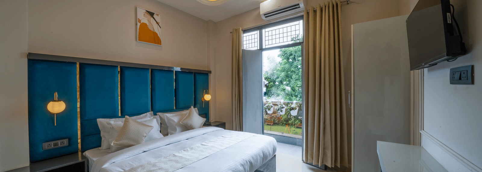 A spacious bedroom featuring a bed with a teal tufted headboard, large window with sheer curtains, a desk, a ceiling fan, and a television - Perfectstayz Value Aalaya Suites, Haridwar
