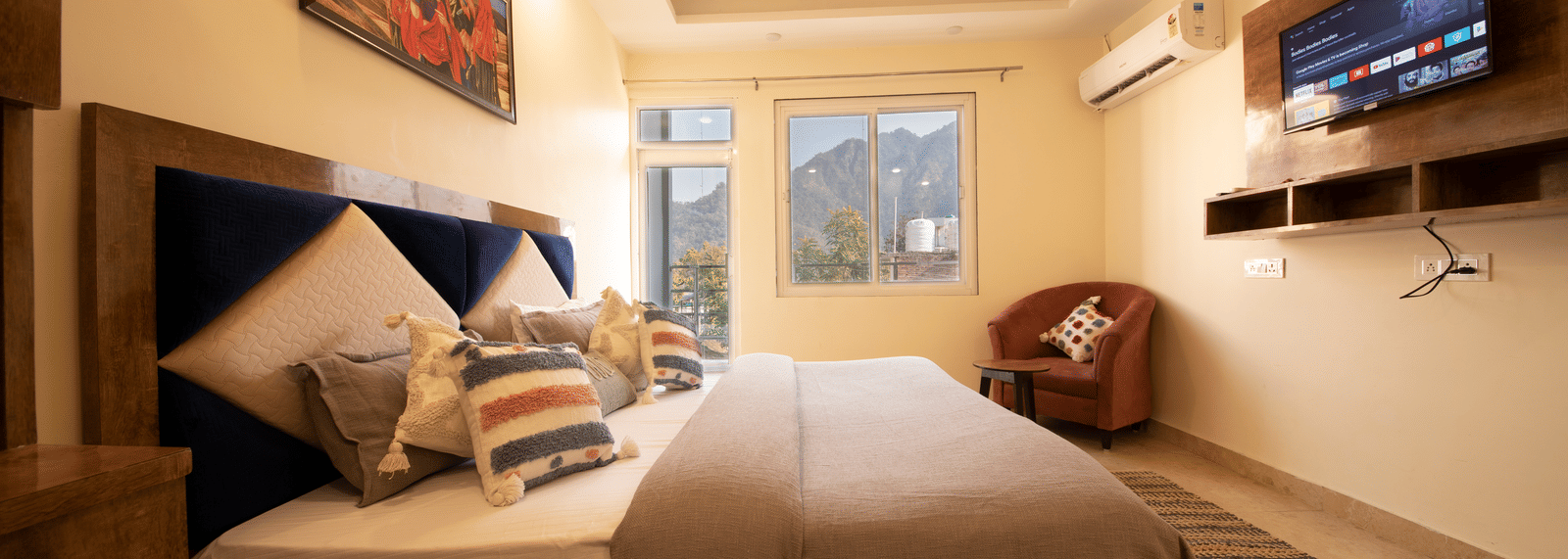 A hotel room, featuring a large grey bed with white linens, blue and orange pillows, a wooden frame, a wall-mounted TV, and a large window with white curtains | Perfectstayz Value Hills, Rishikesh